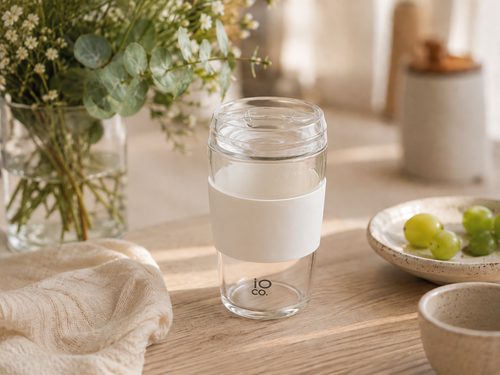 IOco all-glass travel cup in Snow White, with a clear glass lid, matching silicone seal and grip sleeve, shown on a wood dining table with ceramic plates and eucalyptus in a glass vase. Lorikeet Living.