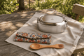 Stainless steel oval bento box with secure lid, placed on a beige cloth on a rustic wooden table outdoors, with a floral fabric utensil pouch and wooden fork