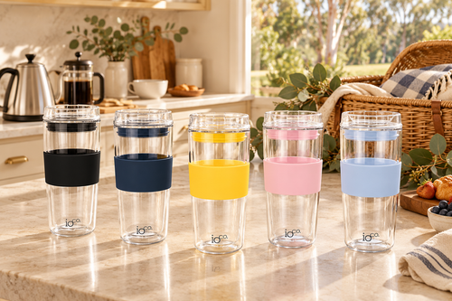 Five IOco all-glass travel cups with black, navy, yellow, pink and pale blue sleeves on a sunlit kitchen bench, ready for a family picnic.
