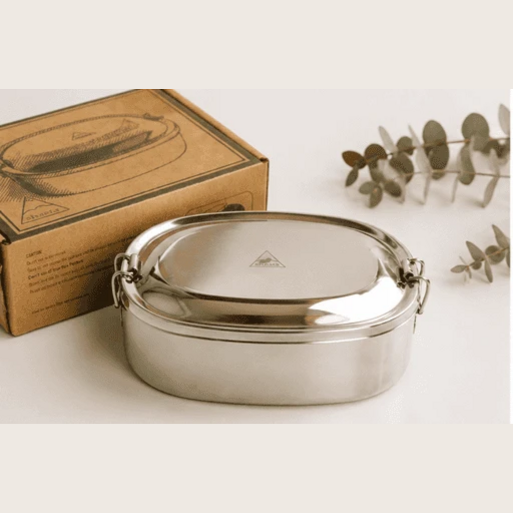 Shasta oval stainless steel bento box with its lid secured, beside its cardboard packaging and eucalyptus sprigs on a neutral background. Lorikeet Living.